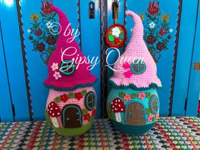 May include: Two crocheted gnome houses, one pink and one blue, with a whimsical design featuring flowers, mushrooms, and windows. The houses are standing in front of a blue door with floral designs.