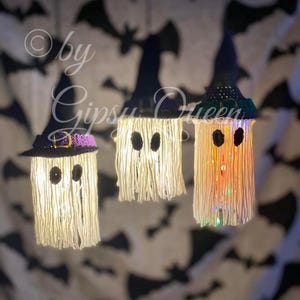 K&ouml;nnte beinhalten: Drei handgefertigte Halloween-Geisterdekorationen. Jeder Geist besteht aus wei&szlig;em Garn mit schwarzen Augen und einem Hexenhut. Die H&uuml;te sind schwarz mit lila und goldenen Akzenten. Der Hintergrund ist ein Stoff mit einem Fledermausmuster.