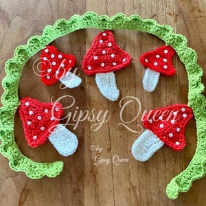 May include: A green crocheted garland with five red and white crocheted mushrooms. The mushrooms have white stems and red caps with white polka dots. The garland is decorated with the text "Gipsy Queen".