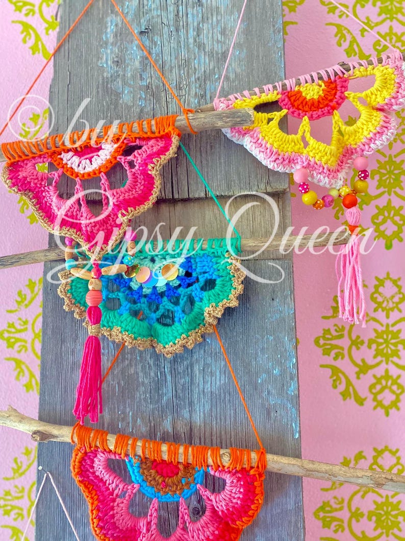 Pode incluir: Tr&ecirc;s tape&ccedil;arias de parede de croch&ecirc; coloridas com franjas, cada uma pendurada em um galho em um fundo de madeira. As tape&ccedil;arias s&atilde;o em tons de rosa, amarelo, laranja, verde e azul.