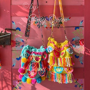 May include: Two colorful crocheted bags with fringe and pom-poms hanging on hooks against a pink wall with floral designs. The bags are made with bright colors like yellow, pink, blue, green, and orange.