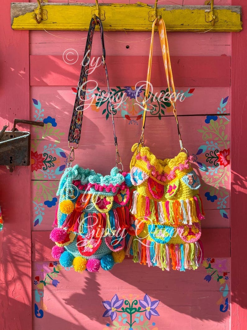 May include: Two colorful crocheted bags with fringe and pom-poms hanging on hooks against a pink wall with floral designs. The bags are made with bright colors like yellow, pink, blue, green, and orange.