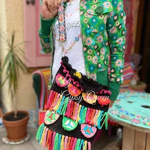 May include: A black crochet bag with colorful fringe and floral accents. The bag has a long strap and is being worn by a person wearing a green floral jacket.