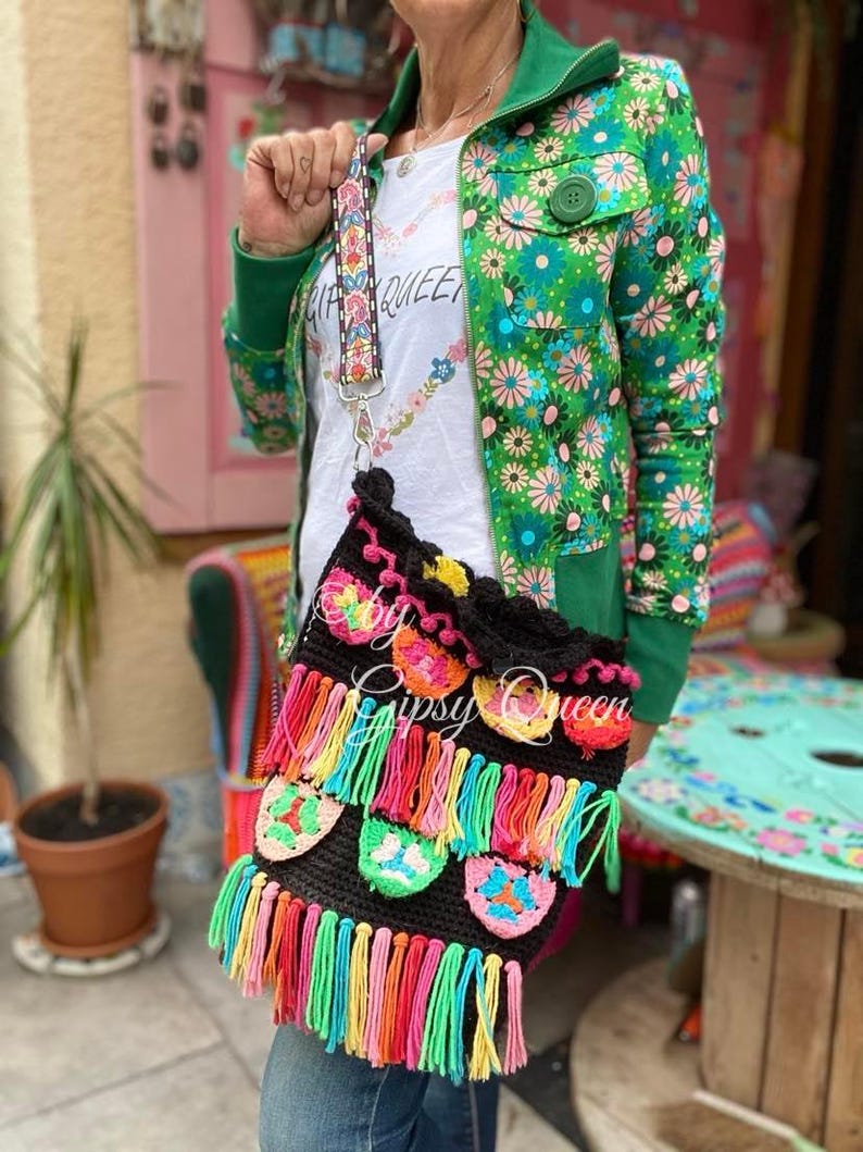 May include: A black crochet bag with colorful fringe and floral accents. The bag has a long strap and is being worn by a person wearing a green floral jacket.