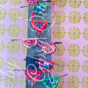 Pode incluir: Uma cole&ccedil;&atilde;o de tape&ccedil;arias de parede de croch&ecirc; coloridas com borlas de contas, exibidas em uma parede de madeira. As tape&ccedil;arias s&atilde;o de v&aacute;rias formas e tamanhos, com uma mistura de cores rosa, verde, azul, laranja e amarelo.