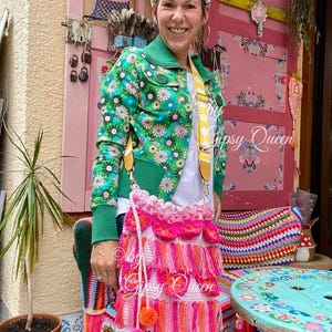 May include: A pink crocheted bag with fringe and pom-poms. The bag is worn over the shoulder of a person wearing a green and floral jacket. The bag is decorated with pink and orange tassels.