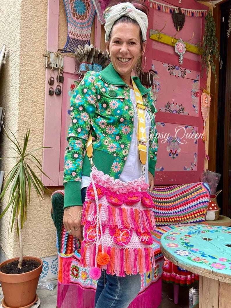 May include: A pink crocheted bag with fringe and pom-poms. The bag is worn over the shoulder of a person wearing a green and floral jacket. The bag is decorated with pink and orange tassels.