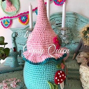 May include: A crocheted gnome house with a pink pointed hat, teal body, and a red and white mushroom appliqu&eacute;. The gnome house is sitting on a teal surface with a crocheted flamingo in the background.