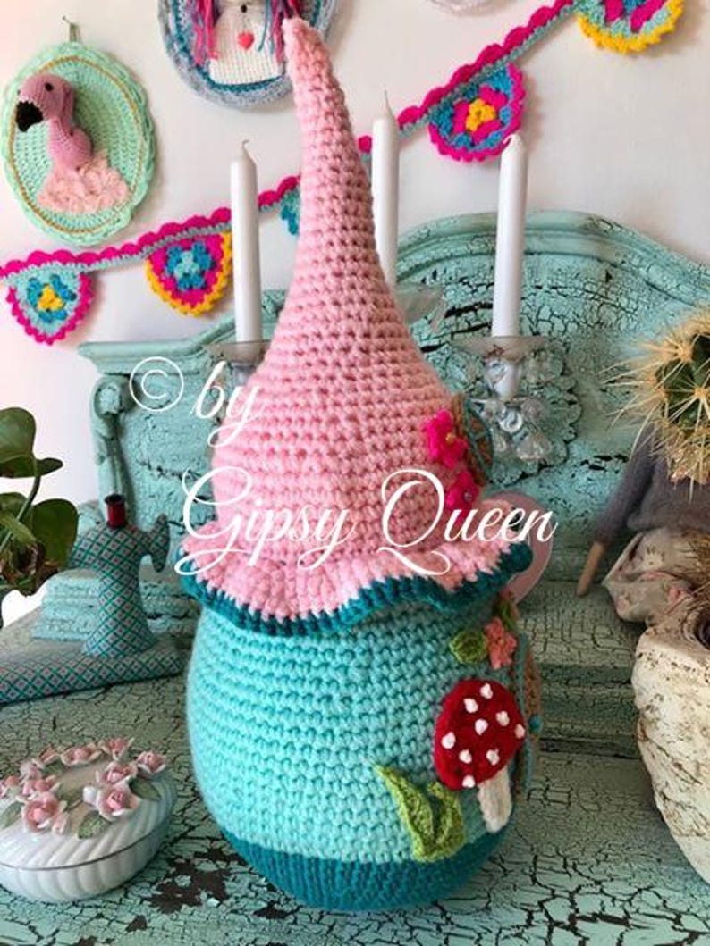 May include: A crocheted gnome house with a pink pointed hat, teal body, and a red and white mushroom appliqu&eacute;. The gnome house is sitting on a teal surface with a crocheted flamingo in the background.