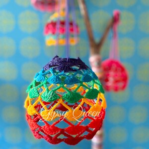 May include: Rainbow crocheted egg ornaments hanging from a branch. The eggs are made with a variety of colors, including red, orange, yellow, green, blue, and purple. The text "By Gipsy Queen" is visible on the bottom of the largest egg.