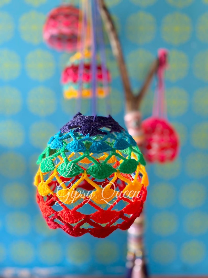 May include: Rainbow crocheted egg ornaments hanging from a branch. The eggs are made with a variety of colors, including red, orange, yellow, green, blue, and purple. The text "By Gipsy Queen" is visible on the bottom of the largest egg.