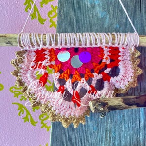 Pode incluir: Uma tape&ccedil;aria de parede de croch&ecirc; rosa, laranja e vermelha com acabamento dourado e lantejoulas iridescentes e roxas. A tape&ccedil;aria est&aacute; presa a um galho.