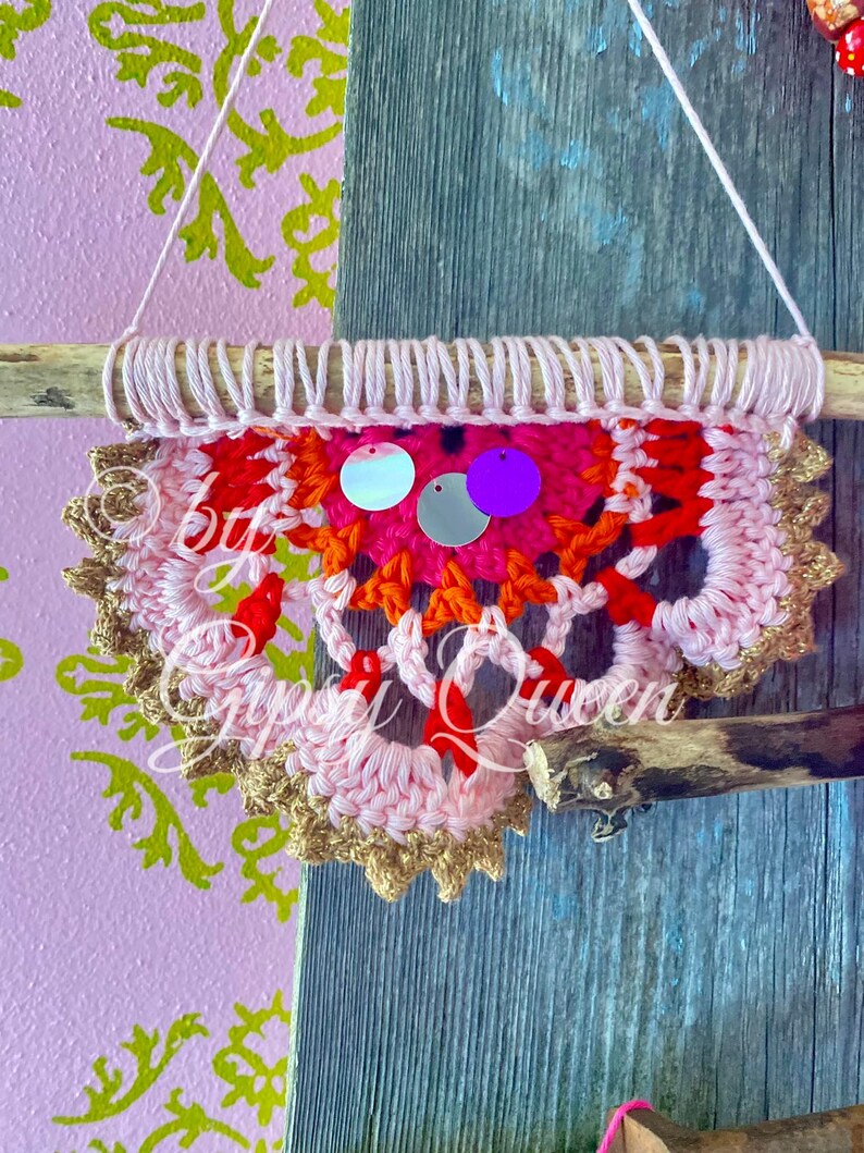 Pode incluir: Uma tape&ccedil;aria de parede de croch&ecirc; rosa, laranja e vermelha com acabamento dourado e lantejoulas iridescentes e roxas. A tape&ccedil;aria est&aacute; presa a um galho.