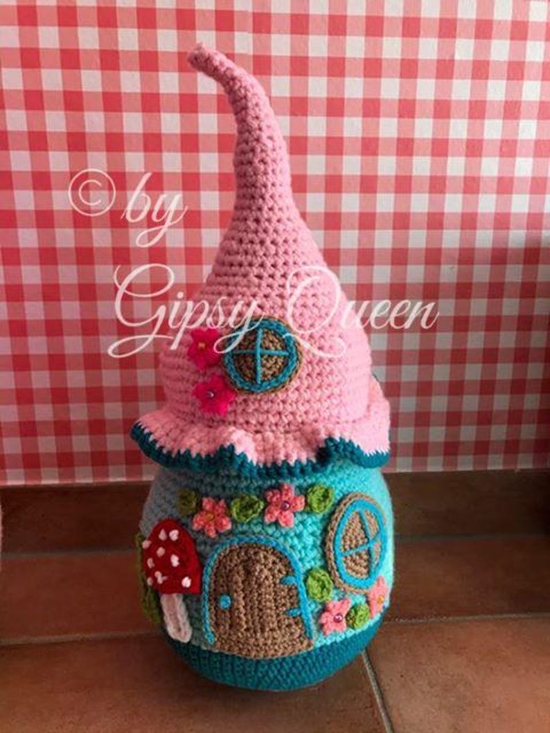 May include: A crocheted gnome house with a pink roof, blue and green details, and a mushroom door. The house is decorated with flowers and a window. The text "by Gipsy Queen" is visible on the image.
