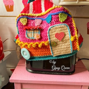 May include: A colorful, crocheted toaster cover shaped like a whimsical house. The cover features a pink body, red roof with white stripes, and various decorative elements in yellow, blue, and green. The door has a red heart. The text "&copy; by Gipsy Queen" is visible.