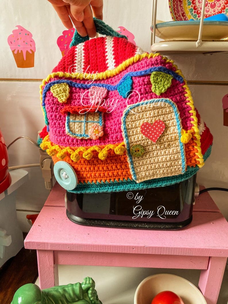 May include: A colorful, crocheted toaster cover shaped like a whimsical house. The cover features a pink body, red roof with white stripes, and various decorative elements in yellow, blue, and green. The door has a red heart. The text "&copy; by Gipsy Queen" is visible.