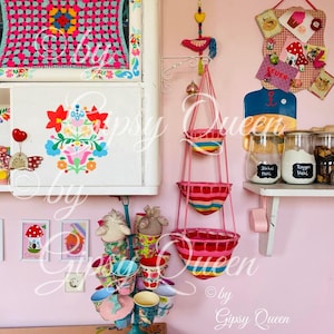 May include: A pink and white kitchen with a colorful crocheted wall hanging, a hanging basket with colorful stripes, and a set of colorful mugs on a stand.