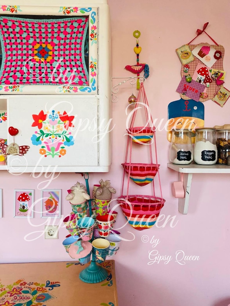 May include: A pink and white kitchen with a colorful crocheted wall hanging, a hanging basket with colorful stripes, and a set of colorful mugs on a stand.