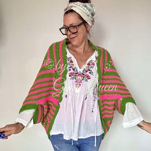 Puede incluir: Un cárdigan de ganchillo a rayas verdes y rosas. El cárdigan tiene un cuello ancho y mangas. La modelo lleva un top blanco bordado y vaqueros azules. El texto "by G Queen" es visible en el cárdigan.