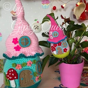 May include: Two crocheted gnome houses, one pink and one green, with a mushroom and flower accents. The houses are sitting on a table with a pink flower pot and a plant.