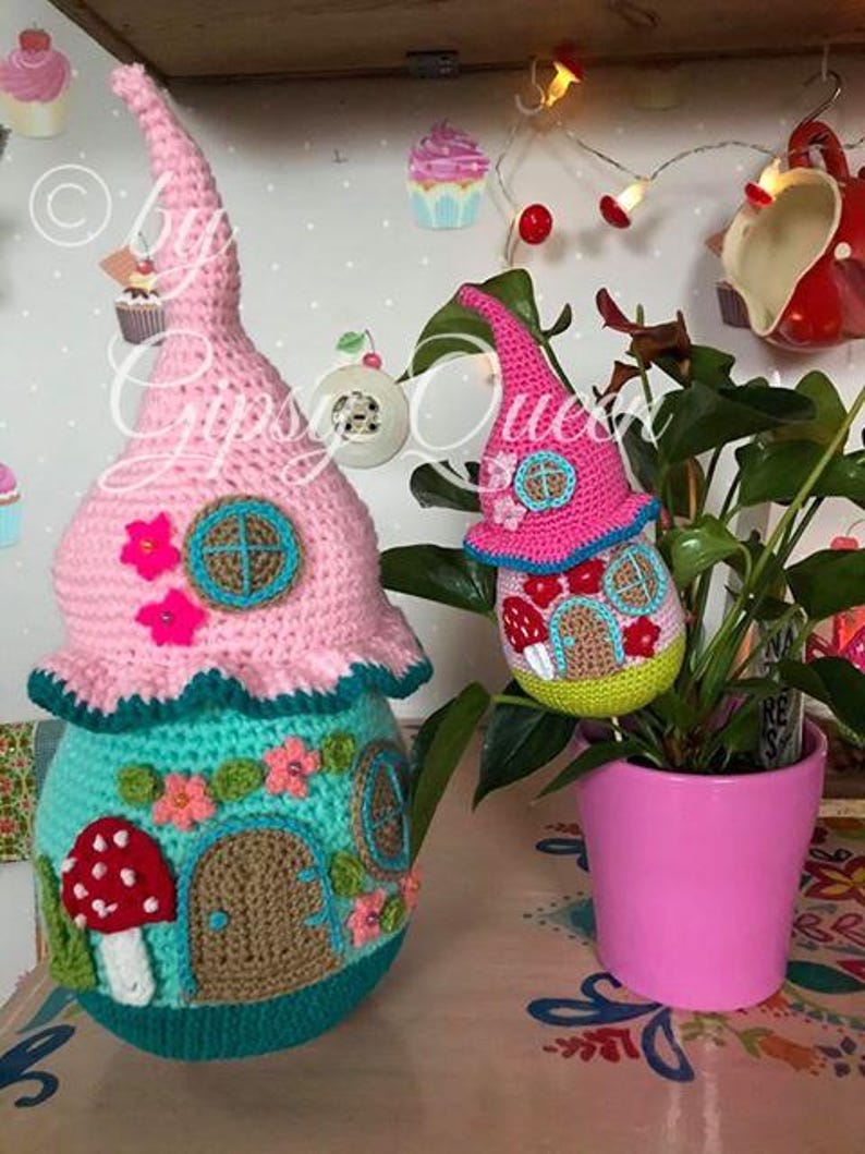 May include: Two crocheted gnome houses, one pink and one green, with a mushroom and flower accents. The houses are sitting on a table with a pink flower pot and a plant.