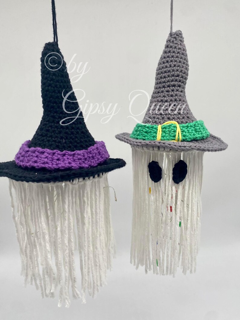K&ouml;nnte beinhalten: Zwei handgefertigte Halloween-Dekorationen aus H&auml;kelarbeit. Eine ist ein schwarzer Hexenhut mit lila Krempe, die andere ein grauer Hexenhut mit gr&uuml;ner Krempe. Beide haben wei&szlig;e Garnstr&auml;hnen, die herunterh&auml;ngen. Der Text "by Gipsy Queen" ist sichtbar.