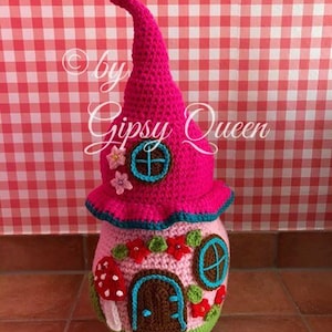 May include: A pink crocheted gnome house with a pointed roof, brown door, and blue windows. The house is decorated with green leaves, red flowers, and a mushroom.