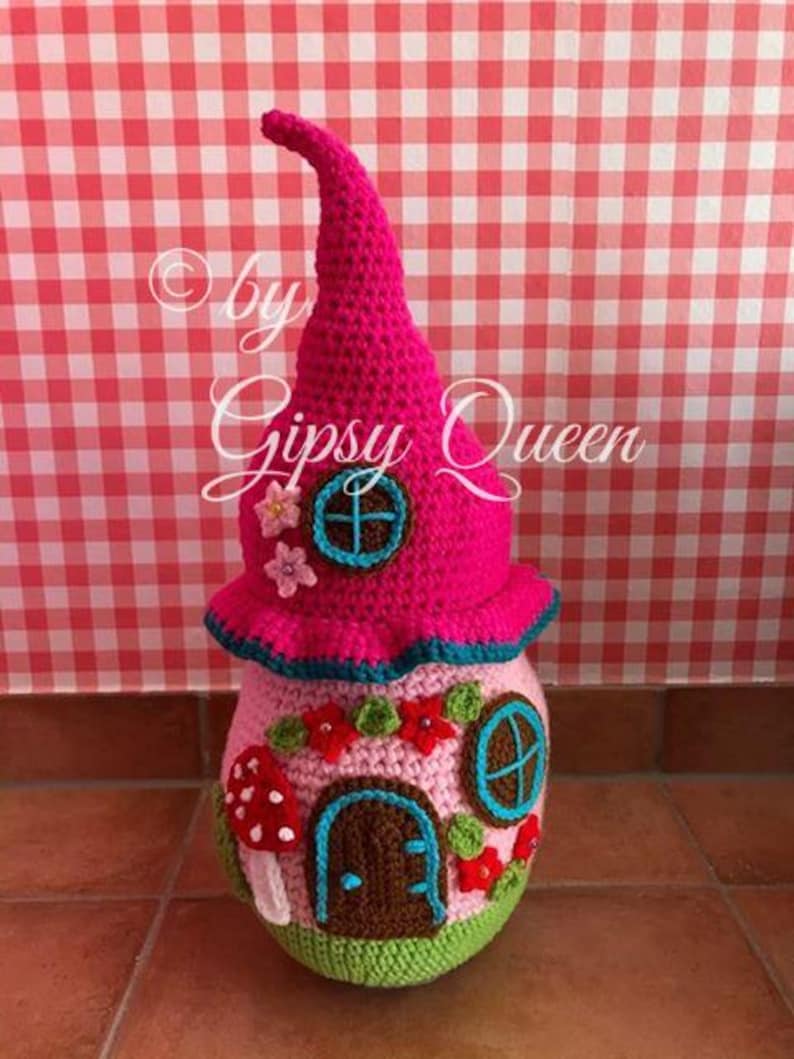 May include: A pink crocheted gnome house with a pointed roof, brown door, and blue windows. The house is decorated with green leaves, red flowers, and a mushroom.