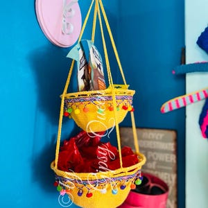 May include: Two yellow crocheted hanging baskets with colorful pom-poms around the rim. The baskets are hanging from a white metal hook attached to a pink wall.