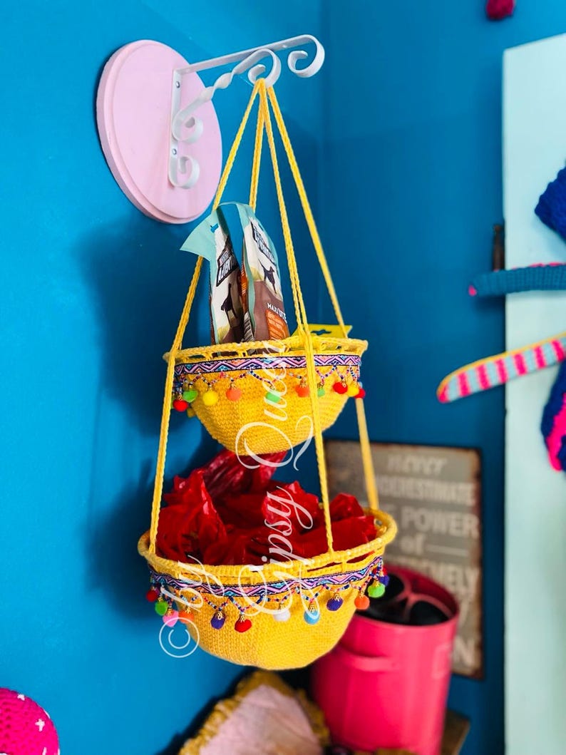 May include: Two yellow crocheted hanging baskets with colorful pom-poms around the rim. The baskets are hanging from a white metal hook attached to a pink wall.