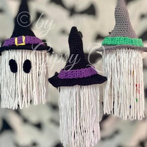 K&ouml;nnte beinhalten: Drei handgefertigte Halloween-Dekorationen. Jede hat einen wei&szlig;en, gefransten K&ouml;rper und einen geh&auml;kelten Hexenhut in Schwarz, Lila und Grau. Ein Hut hat eine gelbe Schnalle. Der Hintergrund hat ein Muster aus schwarzen Flederm&auml;usen.