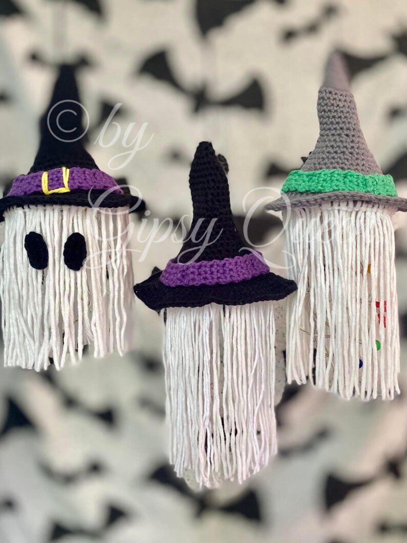 K&ouml;nnte beinhalten: Drei handgefertigte Halloween-Dekorationen. Jede hat einen wei&szlig;en, gefransten K&ouml;rper und einen geh&auml;kelten Hexenhut in Schwarz, Lila und Grau. Ein Hut hat eine gelbe Schnalle. Der Hintergrund hat ein Muster aus schwarzen Flederm&auml;usen.