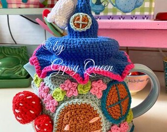 Pode incluir: Uma chaleira de crochê em forma de casa com telhado azul, flores rosa e verdes e uma porta marrom. A chaleira tem uma alça azul e uma campainha branca no topo. O texto "by Gipsy Queen" é visível no lado.