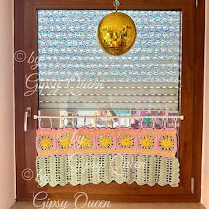 May include: A crocheted window valance with a scalloped bottom edge. The valance is made with pink, yellow, and green yarn and features a repeating pattern of squares with flower designs. The valance is hanging on a window with a disco ball hanging from the ceiling above.