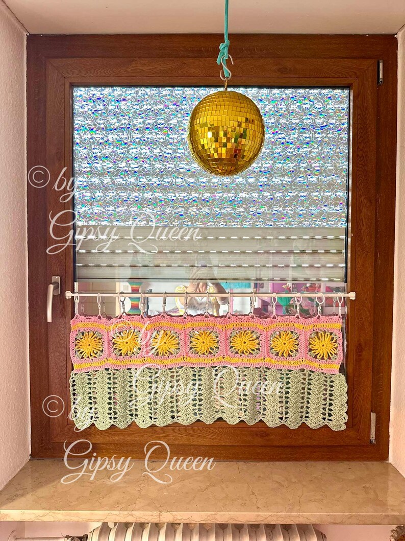 May include: A crocheted window valance with a scalloped bottom edge. The valance is made with pink, yellow, and green yarn and features a repeating pattern of squares with flower designs. The valance is hanging on a window with a disco ball hanging from the ceiling above.