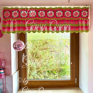 May include: A pink and green crocheted window valance with a scalloped edge. The valance features a repeating pattern of squares with a flower design in the center. The valance is hung on a window with a view of green trees.