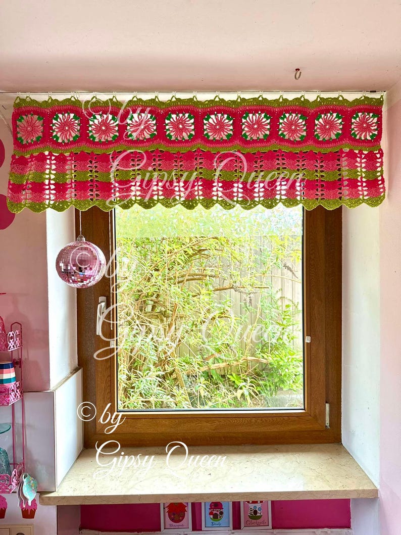 May include: A pink and green crocheted window valance with a scalloped edge. The valance features a repeating pattern of squares with a flower design in the center. The valance is hung on a window with a view of green trees.