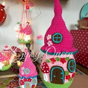 May include: Three crocheted fairy houses in pink, green, and white with a mushroom and flower design. The largest house has a tall pink pointed roof and a brown door. The smallest house is hanging from a string.