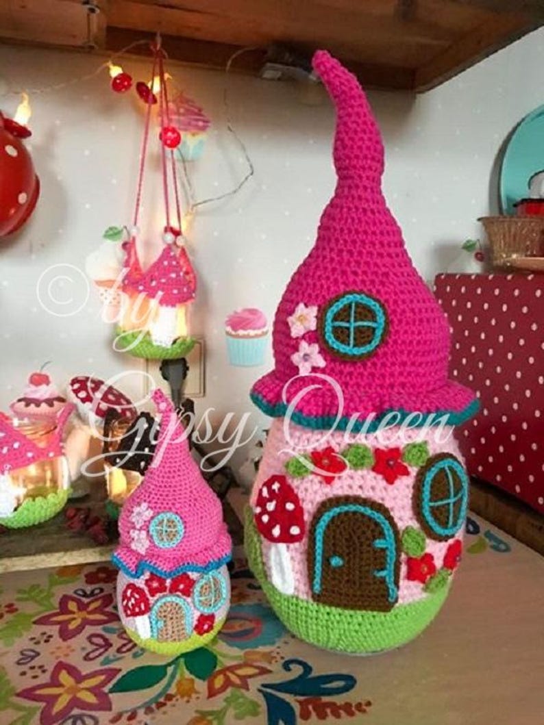 May include: Three crocheted fairy houses in pink, green, and white with a mushroom and flower design. The largest house has a tall pink pointed roof and a brown door. The smallest house is hanging from a string.