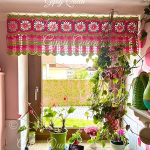 May include: A pink and green crocheted window valance with a scalloped bottom edge. The valance features a repeating pattern of flowers and leaves. The valance is hanging in a window with plants on the windowsill.