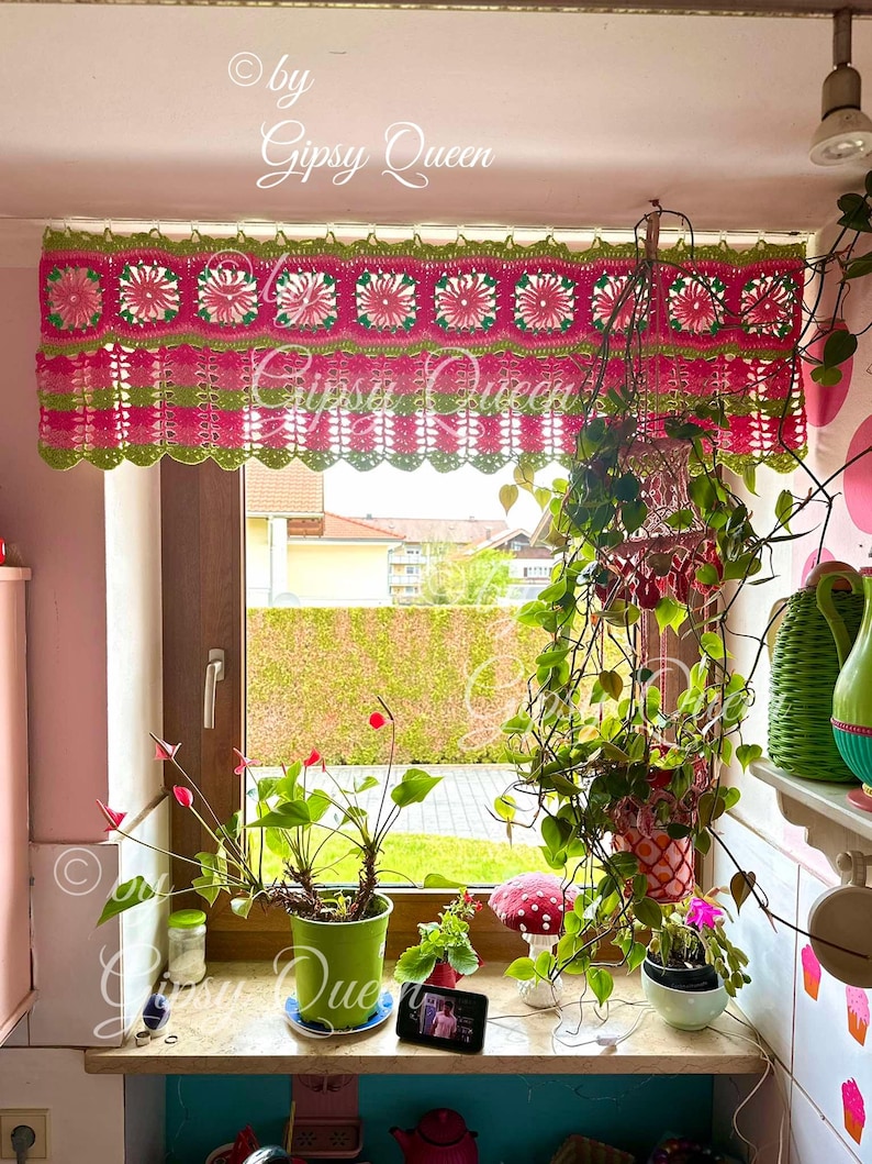 May include: A pink and green crocheted window valance with a scalloped bottom edge. The valance features a repeating pattern of flowers and leaves. The valance is hanging in a window with plants on the windowsill.