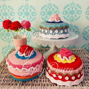 Könnte beinhalten: Drei gehäkelte Kuchen auf einem weißen Kuchenständer und einer Oberfläche aus Weide. Die Kuchen sind mit bunten gehäkelten Blumen und Verzierungen verziert. Der Kuchen auf dem Ständer ist blau mit weißen und goldenen Verzierungen. Der Kuchen links ist rosa mit Regenbogenverzierungen. Der Kuchen rechts ist rot mit weißen und goldenen Verzierungen.