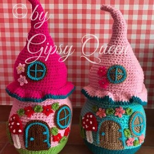 May include: Two crocheted gnome houses, one pink and one blue, with a brown door, a window, and mushroom accents. The houses are sitting on a red and white checkered background.