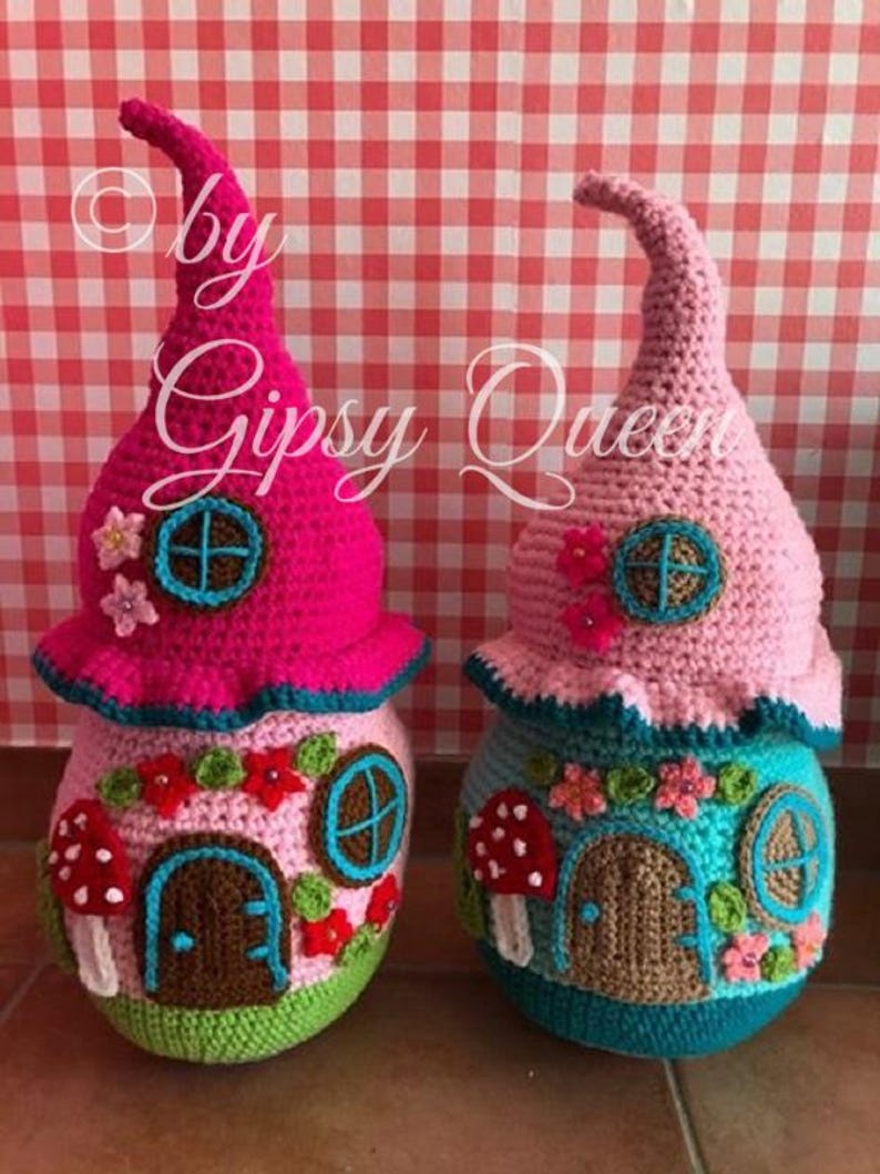 May include: Two crocheted gnome houses, one pink and one blue, with a brown door, a window, and mushroom accents. The houses are sitting on a red and white checkered background.