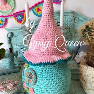 May include: A crocheted gnome house with a pink pointed hat, teal body, and a brown and beige window. The gnome house is sitting on a teal surface with a white and pink floral design.