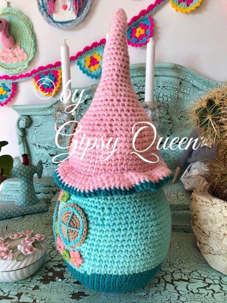 May include: A crocheted gnome house with a pink pointed hat, teal body, and a brown and beige window. The gnome house is sitting on a teal surface with a white and pink floral design.
