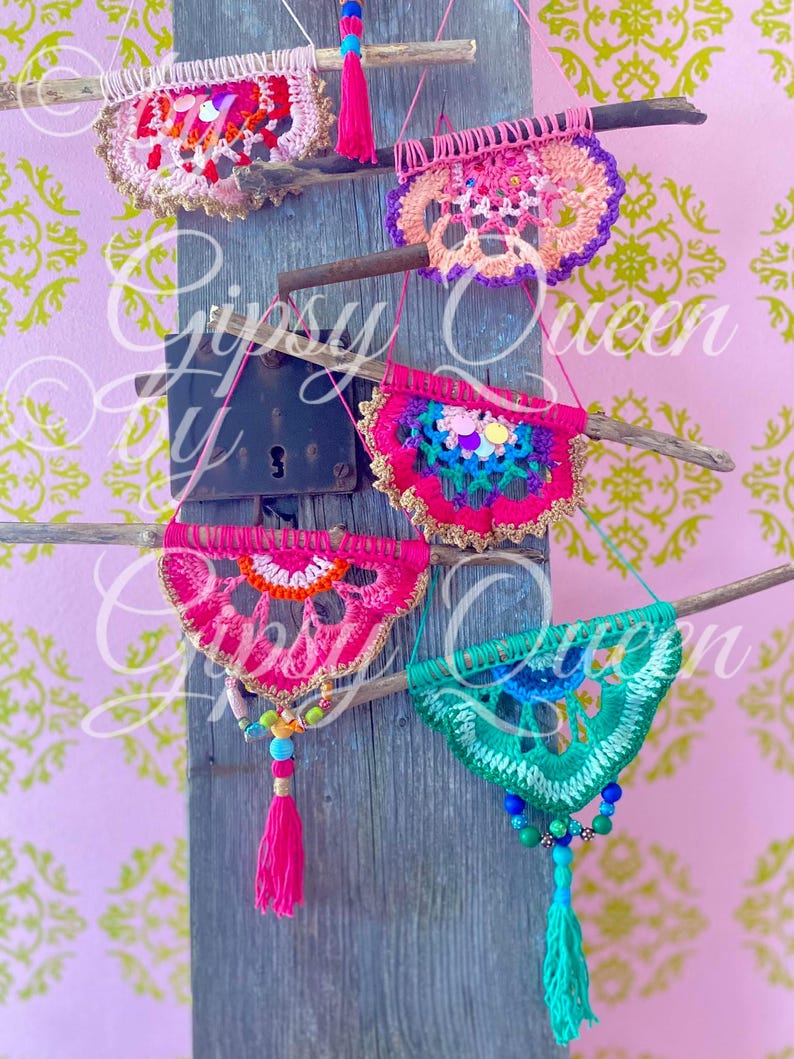 Pode incluir: Cinco tape&ccedil;arias de parede de croch&ecirc; em v&aacute;rias cores, incluindo rosa, verde, azul e laranja, s&atilde;o exibidas em uma superf&iacute;cie de madeira. Cada tape&ccedil;aria apresenta um design circular com uma franja de contas.