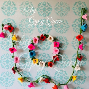 Puede incluir: Una guirnalda y una corona de flores de ganchillo en varios colores, incluyendo rosa, rojo, amarillo, blanco y azul. La guirnalda y la corona están dispuestas sobre una pared azul claro con un patrón repetido. El texto "by Gipsy Queen" es visible.