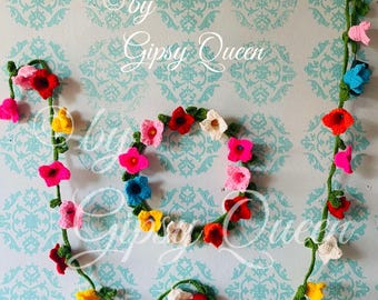 May include: A crocheted floral garland and wreath in various colors, including pink, red, yellow, white, and blue. The garland and wreath are arranged on a light blue wall with a repeating pattern. The text "by Gipsy Queen" is visible.