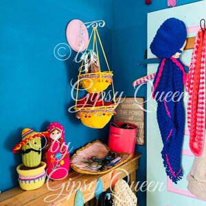 May include: A teal blue wall with a pink and white hanging wall organizer with two yellow crocheted baskets. The wall organizer is hanging from a white metal hook. There is a wooden shelf with a pink and white crocheted doll, a green crocheted cactus, a pink container, and a woven tray with keys and other small items. The shelf is decorated with a sign that reads "Never underestimate the power of extremely off plan".